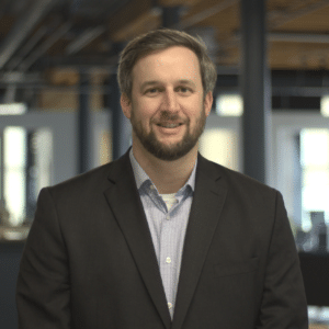 Jared, an Exit Planning Advisor, wearing a dark suit and light checkered shirt, standing in an office environment smiling at the camera.